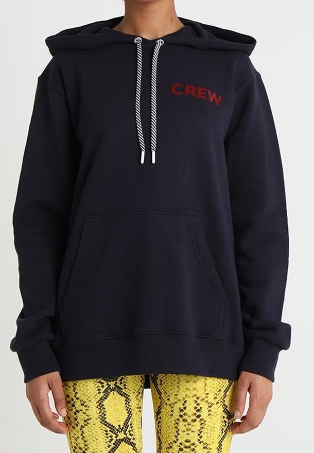 Personne portant un hoodie bleu marine avec "CREW" en lettres rouges et des cordons blancs, assorti à un pantalon jaune à motif serpent.