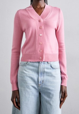Kobieta ubrana w różowy kardigan zapinany na guziki z prążkowanymi mankietami oraz jasnoniebieskie, wysokowalne dżinsy na tle jednolitego tła.