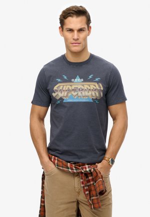 T-shirt bleu marine avec un graphique audacieux "SUPERDRY" en beige et turquoise. Manches courtes, col rond, porté avec une chemise à carreaux nouée autour de la taille.