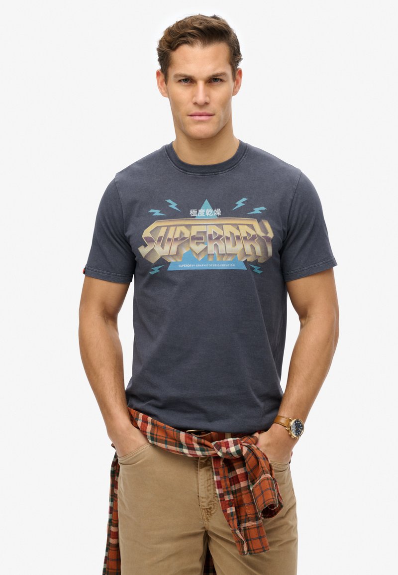 Camiseta de color navy con un atrevido gráfico "SUPERDRY" en beige y turquesa. Mangas cortas, cuello redondo, combinada con una camisa de cuadros atada a la cintura.