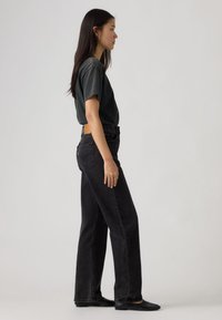 Νεαρή γυναίκα όρθια πλάγια, φοράει άνετο μαύρο t-shirt, ψηλόμεσο μαύρο τζιν και μαύρα φλατ παπούτσια, με φόντο ένα απλό υπόβαθρο.