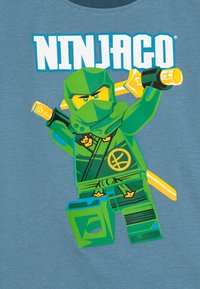 Green Ninjago character graphic on a blue shirt, featuring sword, tactical gear, and a circular emblem. Smooth fabric texture, short sleeves.