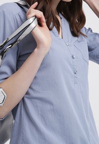 Chemise bleu clair à boutons avec une petite poche poitrine, présentant des rayures verticales et une coupe décontractée. Sangle de sac à main argentée visible.