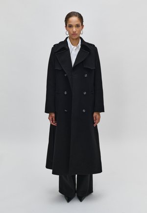 Manteau trench en laine noire avec un design croisé, grands revers, poches avant et détails en boutons. Porté sur une chemise blanche et un pantalon sombre.