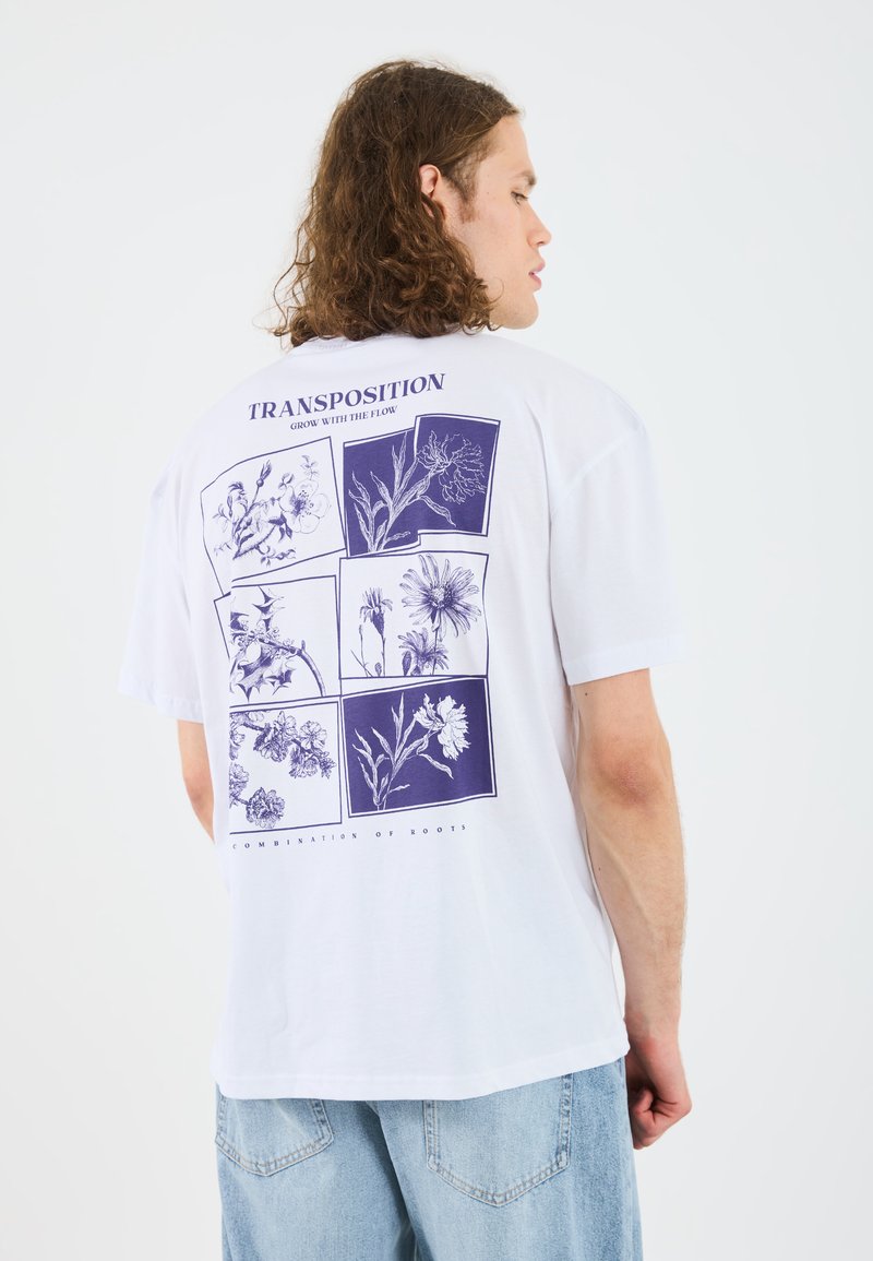 Hvit T-skjorte med lilla botaniske illustrasjoner på baksiden, med teksten "TRANSPOSITION" og "GROW WITH THE FLOW." Korte ermer, avslappet passform.