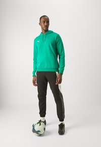 Sweatshirt verde com bolso canguru, combinado com calças de fato pretas com painel lateral branco. Sapatos de futebol pretos e uma bola de futebol multicolorida.