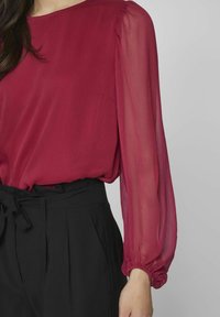 Donna che indossa una blusa bordeaux trasparente con maniche lunghe, infilata in pantaloni neri a vita alta con una cintura annodata in vita.