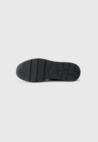 Black rubber shoe sole featuring a zigzag tread pattern and indentations for grip, with a smooth surface and rounded toe shape.