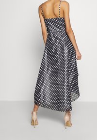 Kvinna i en svart satin midi-klänning med vita prickar och tunna band, tillsammans med beige högklackade sandaler, sedd bakifrån.