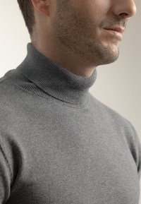 Hombre con una ligera barba de varios días que lleva un suéter de cuello alto de canalé gris, mostrado desde el cuello hasta los hombros contra un fondo sencillo.