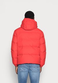 Rød pufferjakke med hætte, der har quiltede paneler og elastiske manchetter, parret med blå jeans. Stoffet ser glat og isoleret ud.