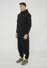 Sweat à capuche zippé noir avec cordon, poches avant, associé à un jogger fuselé assorti. Porté avec des baskets multicolores, présentant des accents gris et rouge.