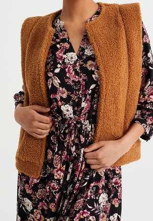 Vrouw draagt een bruine gestructureerde mouwloze vest over een zwarte bloemenjurk met een trekkoord in de taille, zij past het vest aan met beide handen.