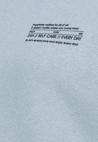 Hellblauer Stoff mit glatter Textur und schwarzem Text, der Selbstfürsorge und Glück betont. Der Text enthält verschiedene Phrasen und Symbole.