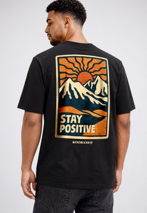 Man wearing black t-shirt with graphic of orange sun, snow-capped mountains, and text "STAY POSITIVE" on back.