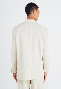 Blazer de color beig claro con un diseño estructurado, cierre de un solo botón y mangas largas. La tela parece suave con un detalle mínimo.
