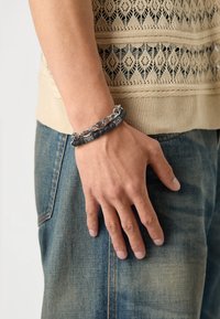 Bracelet en pierre de lave noire et chaîne en argent, avec des perles rondes, des maillons et des accents gravés. Porté sur une main contre un tissu en denim.