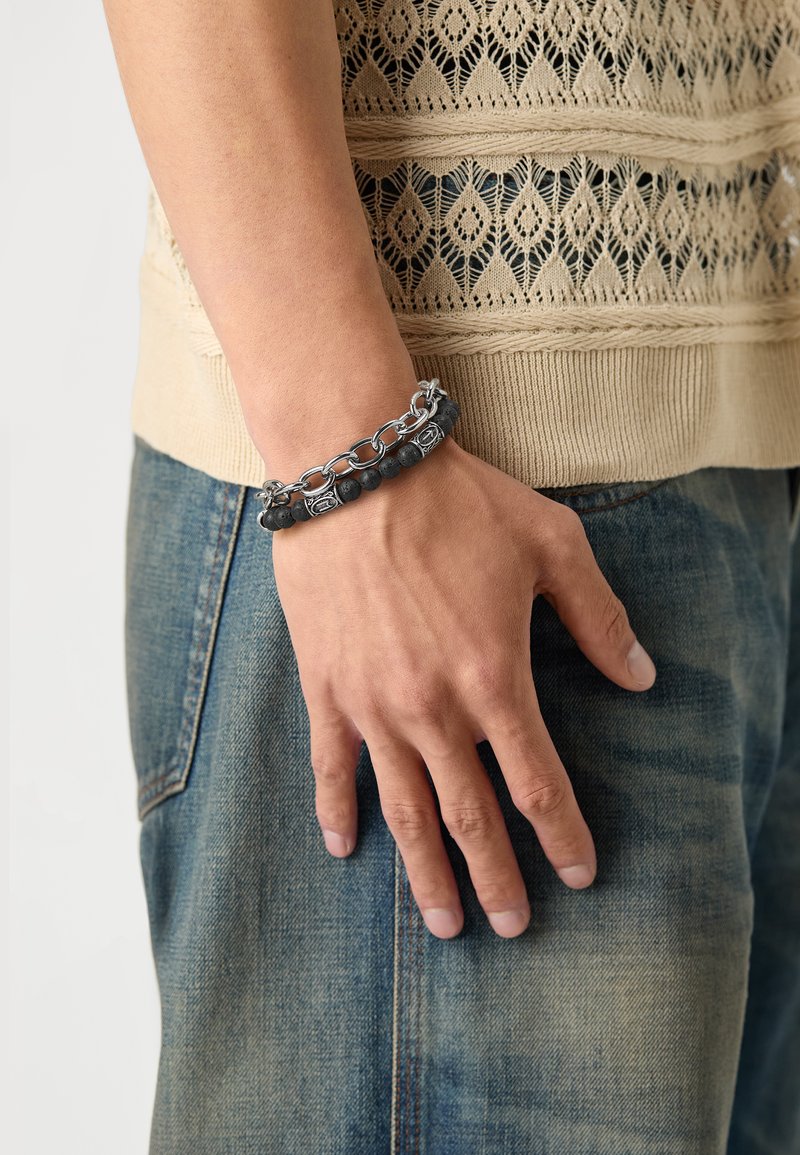 Bracelet en pierre de lave noire et chaîne en argent, avec des perles rondes, des maillons et des accents gravés. Porté sur une main contre un tissu en denim.