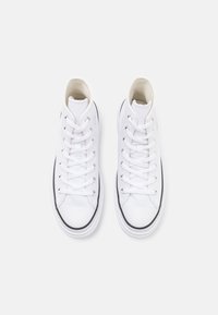 Paire de baskets montantes en toile blanche avec lacets blancs et bordure noire autour des semelles en caoutchouc sur un fond clair.