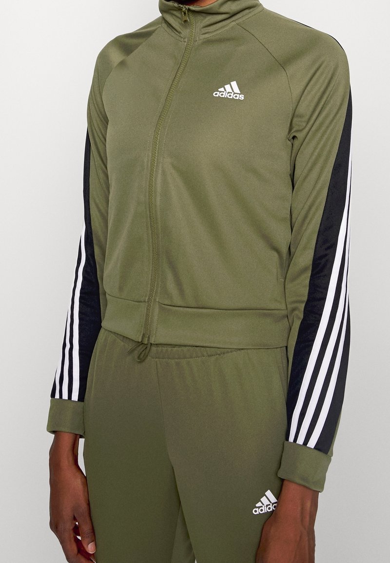 Personne portant un survêtement Adidas vert olive avec des manches rayées noires et blanches et le logo Adidas sur la poitrine et le pantalon.