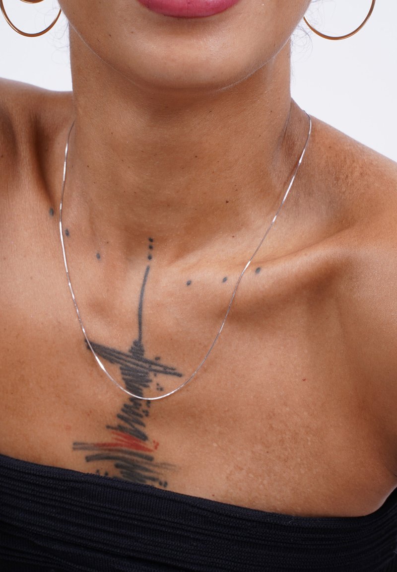 Eine silberne Halskette liegt auf einem Schlüsselbein und wird von filigranen Tattoos begleitet. Der Hautton ist warm, mit einer glatten Textur vor einem dunklen Oberteil.