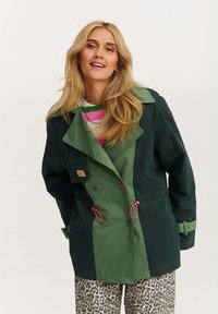 Zelená a tmavě zelená voděodolná bunda s širokým límcem, růžovými akcenty a knoflíky; má dvouřadé zapínání a stahovací pas.