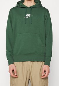 Grön bomullshoodie med framficka, huva med dragsko och vit Nike-logotyp. Har ribbade mudd och nederkant med en jämn textur.