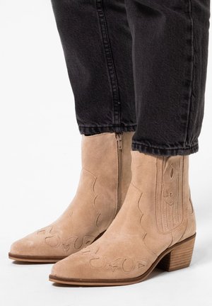 Beige suede ankle boots with pointed toes, decorative stitching, and a low stacked heel, featuring a side zipper for easy wear.