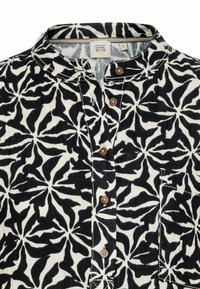 Sort og hvid mønstret skjorte med høj krave, der har blomsterdesigns, brune knapper og en enkelt brystlomme. Lavet af bomuldsstof.