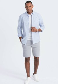 Hellblau-weiße gestreifte Hemdbluse über einem weißen T-Shirt, kombiniert mit hellgrauen Shorts und weißen Turnschuhen. Glatter Stoff und lässige Passform.
