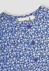 Robe florale bleue en tissu léger, col rond et deux boutons violets à l'avant, avec des motifs de fleurs blanches sur l'ensemble.
