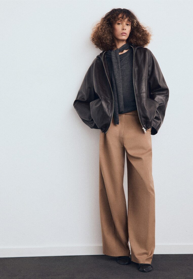 Chaqueta bomber de cuero marrón sobre una camiseta gris oscura, combinada con pantalones anchos beige. Tejido texturizado, ajuste relajado y hardware mínimo.