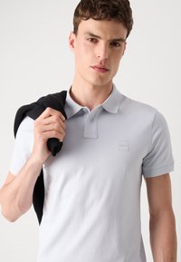 Ανοιχτό γκρι πόλο μπλούζα με κοντά μανίκια, πλάκα με δύο κουμπιά και κεντημένο λογότυπο στο στήθος, συνδυασμένο με έναν μαύρο πλεκτό σκέπασμα στους ώμους.