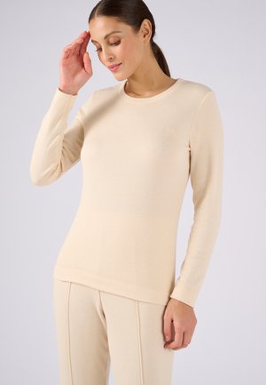 Femme portant un haut à manches longues côtelé beige et un pantalon assorti, la main droite proche de la tête et les yeux fermés.