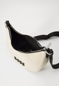 White canvas crossbody bag with black accents, zipper closure, and logo detail on the front. Interior includes a pocket for organization.
