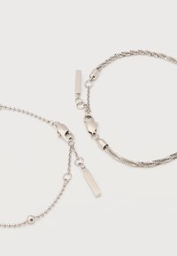 Δύο ασημένια βραχιόλια αλυσίδας με κούμπωμα στυλ αστακού και μικρές ορθογώνιες ετικέτες σε λευκό φόντο.