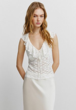 Jeune femme aux longs cheveux ondulés portant un haut blanc en dentelle sans manches avec des volants et une jupe blanche sur un fond uni.