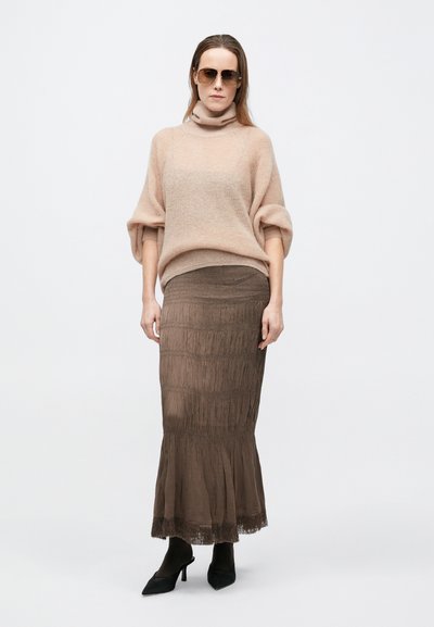 Világos rózsaszín kötött pulóver magas nyakkal, hosszú barna rojtos szoknyával és fekete hegyes orrú bokacsizmával párosítva. A modell napszemüveget visel.