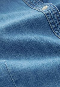 Hellblauer Denimstoff mit einer glatten Textur, der subtile Farbvariationen und eine schwache vertikale Maserung aufweist. Enthält einen Knopf und Nähdetails.
