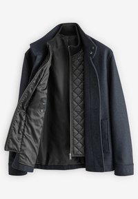 Marineblauer Wollmischmantel mit gefüttertem schwarzen Innenfutter. Verfügt über einen Stehkragen, vordere Knöpfe und Seitentaschen. Innen befindet sich eine Taschen für das Etikett.