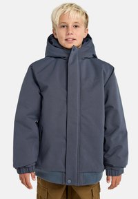 Ragazzo biondo che indossa una giacca invernale blu con cappuccio e tasche con zip, insieme a pantaloni cargo marroni, in piedi contro uno sfondo neutro.