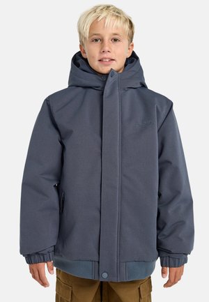 Ragazzo biondo che indossa una giacca invernale blu con cappuccio e tasche con zip, insieme a pantaloni cargo marroni, in piedi contro uno sfondo neutro.