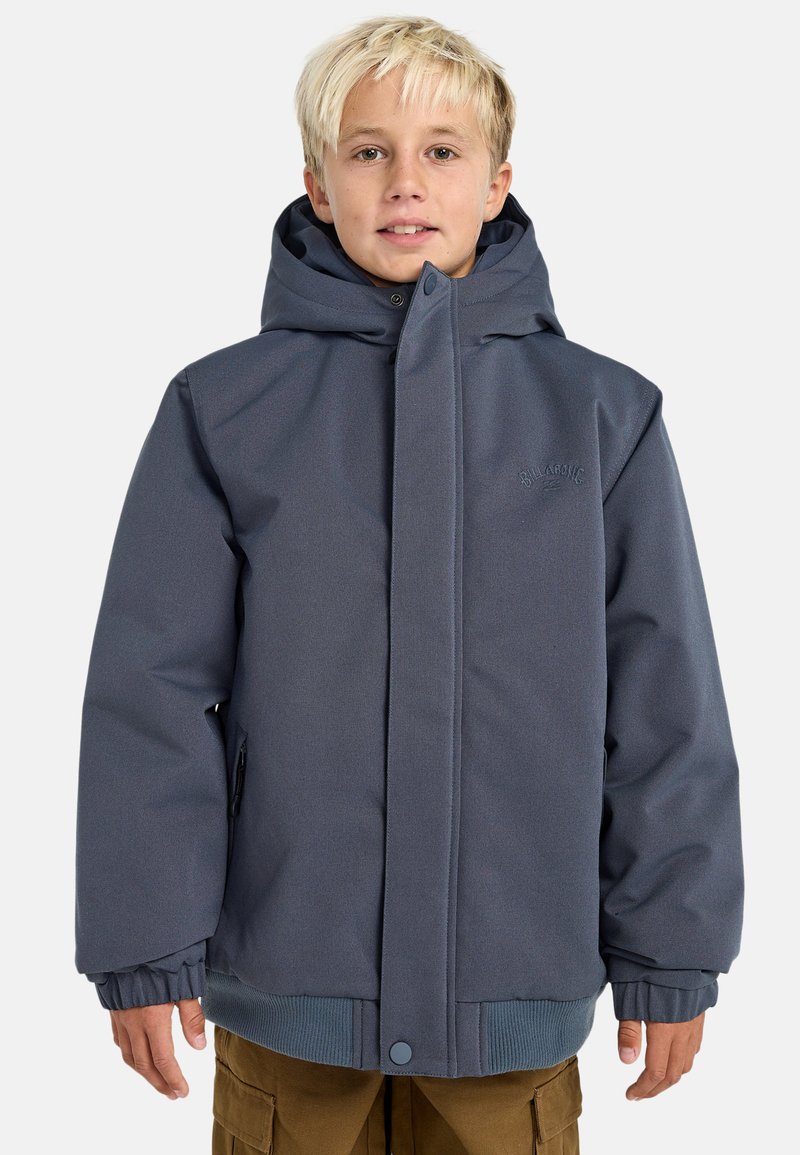 Ragazzo biondo che indossa una giacca invernale blu con cappuccio e tasche con zip, insieme a pantaloni cargo marroni, in piedi contro uno sfondo neutro.