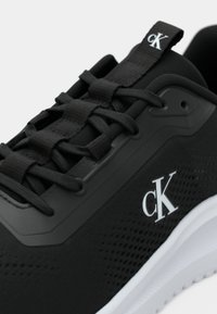 Sapatilha preta com atacadores pretos, logotipo branco "cK" na lateral, aba na língua e sola texturizada branca.