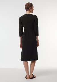 Vestido negro hasta la rodilla con mangas de tres cuartos y una silueta ajustada. Cuenta con un discreto cierre en la parte posterior y pliegues en la cintura.