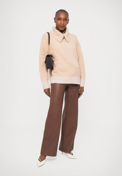 Maglione beige morbido con colletto alto, design a mezza zip, abbinato a pantaloni in ecopelle marroni a gamba larga e tacchi a punta bianchi. Inclusa borsa nera.