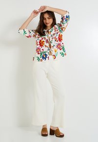 Chaqueta blanca de botón con patrón floral y acentos en rojo, amarillo y verde; combinada con pantalones acampanados blancos y mules de tacón marrones.