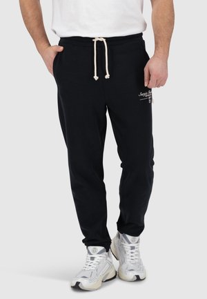 Schwarze Baumwoll-Sweatpants mit einem Kordelzugbund, Seitentaschen und einem Logo-Print am linken Bein. Kombiniert mit weißen Turnschuhen.