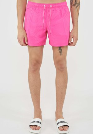Homme portant un short de bain rose vif et des sandales blanches Emporio Armani, avec des tatouages sur l'avant-bras et la cuisse, debout devant un fond blanc.