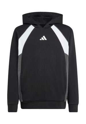 Dreng iført sort og hvid Adidas-hoodie med hænderne i lommerne, smilende mod en ensfarvet lys baggrund.